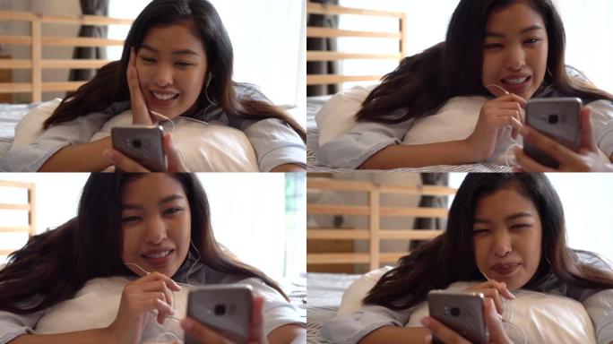 穿着休闲服装的快乐年轻亚洲女孩的肖像躺在床上，一边用智能手机打视频电话，在家里的卧室里挥手。视频会议
