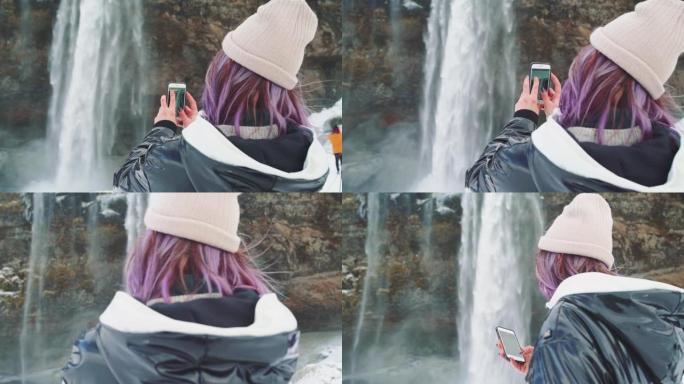 美丽的年轻女子在冰岛的一个瀑布附近，在电话上拍照，欢欣鼓舞并自拍