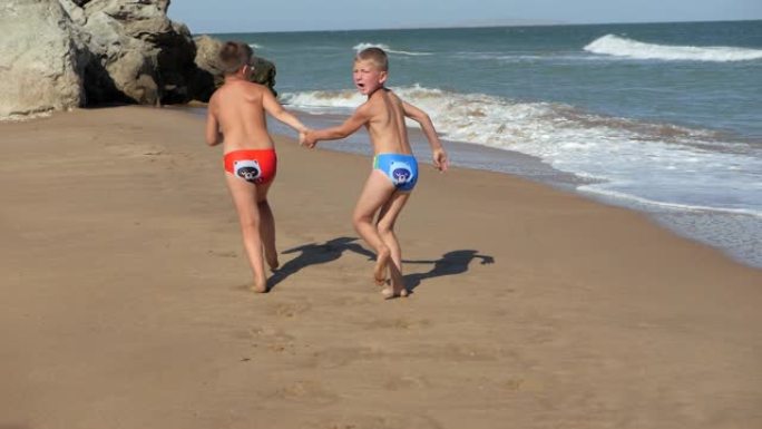 孩子们沿着海岸线奔跑。海浪冲过海滩。男孩们手牵手沿着海滩奔跑。在海边其他地方戴眼镜的孩子。男孩跑着又