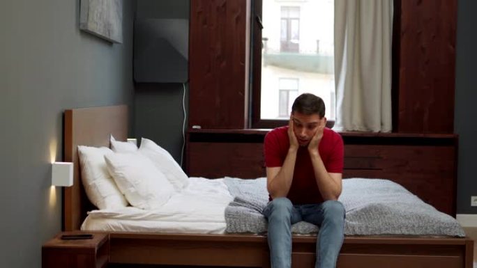沮丧的年轻人坐在家里或酒店房间的床边，焦虑地揉手。紧张的人看着放在床头柜上的手机等待上瘾的电话