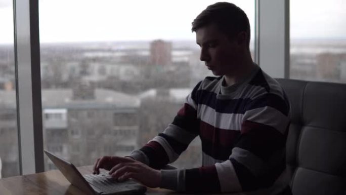 一个年轻人带着一台笔记本电脑坐在一张桌子旁。一名男子坐在全景窗边的咖啡馆里，带着一台小笔记本电脑写信