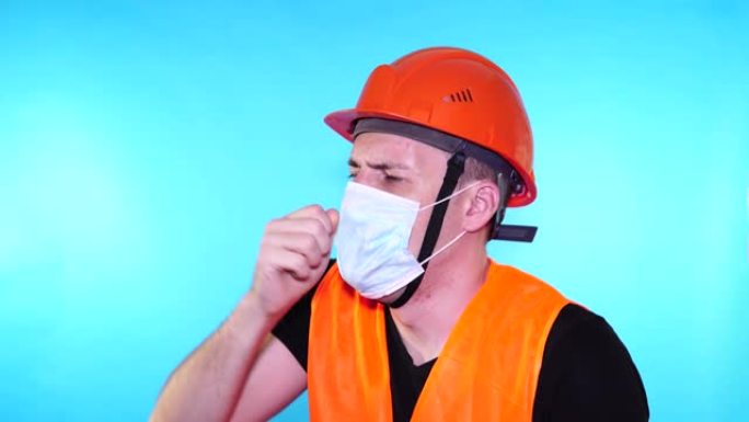 穿着工作服和医用口罩的男性建筑工人在蓝色背景上咳嗽。感染威胁的概念