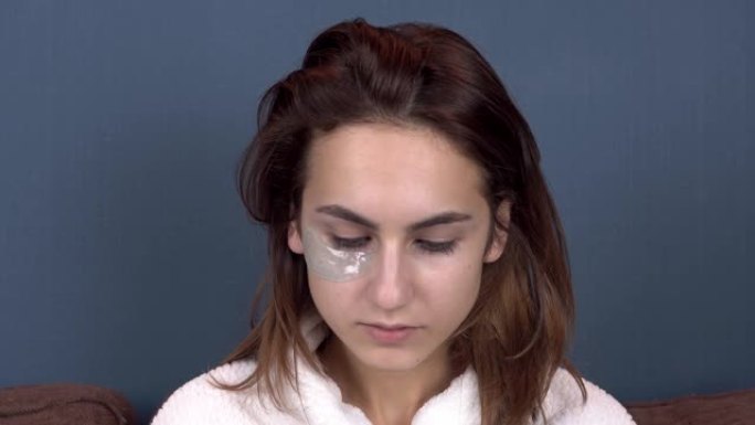 年轻女子在脸上贴了补丁。蓝色水凝胶贴片，用于恢复面部皮肤的活力。坐在白大褂里的女孩。特写