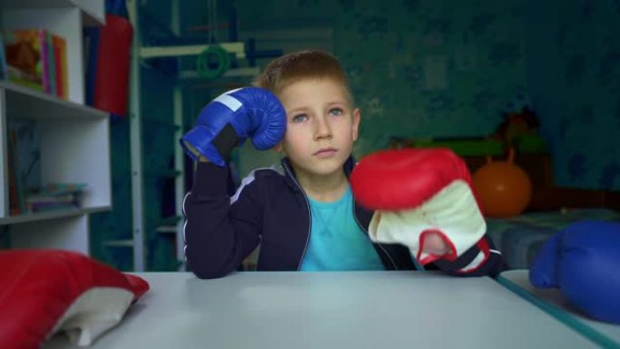 这个男孩梦想和朋友一起玩盒子。戴拳击手套的悲伤男孩坐在桌子旁。