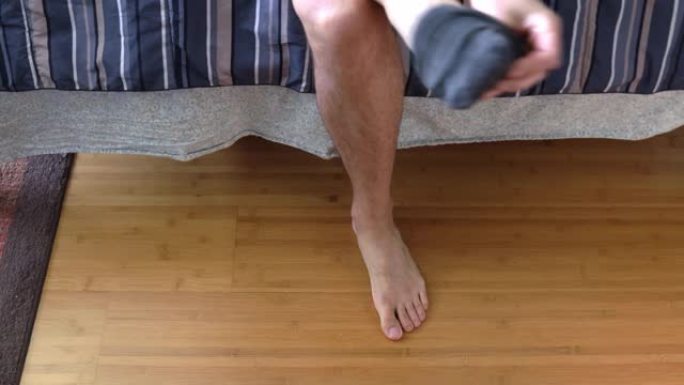 一个白人男性模特的特写镜头，坐在床边，从他的黑色脚踝袜子中换下来，穿上一些破旧的棕色针织拖鞋，然后走