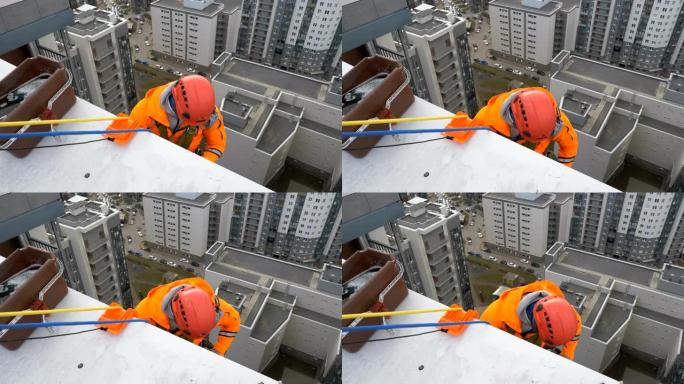 穿着橙色西装和头盔的工业登山者准备从多层建筑的屋顶下降以进行玻璃清洗