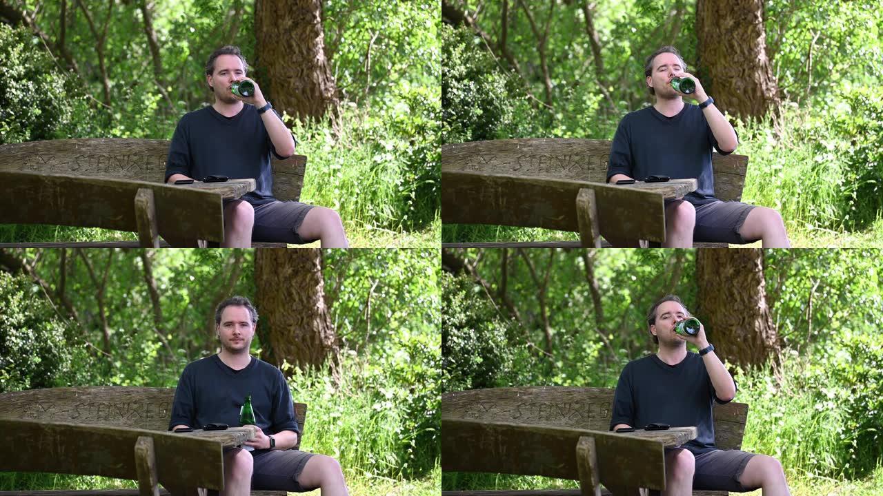 一名男子坐在大自然的木凳上，享受夏日，用绿色玻璃瓶喝水