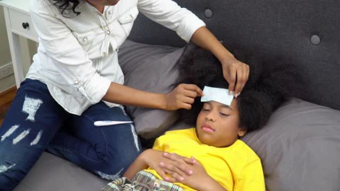 4k非裔美国母亲将发热贴片放在疾病女儿头上，并使用数字口腔温度计检查温度。可爱的小女孩躺在床上感冒和
