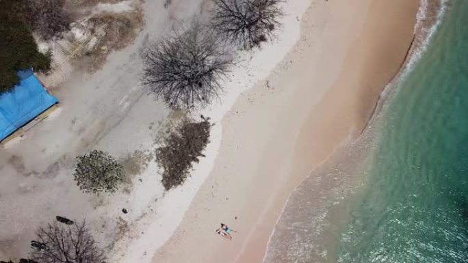 一对夫妇躺在印度尼西亚龙目岛粉红海滩海滨的沙滩上玩无人机。波浪轻轻地冲刷着海岸。他们在晒黑。快乐和粗