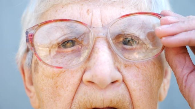 沉思的眼镜中奶奶的细节肖像。关闭女性养老金领取者的皱纹脸，看着相机并调整眼镜。奶奶悲伤的表情。慢动作