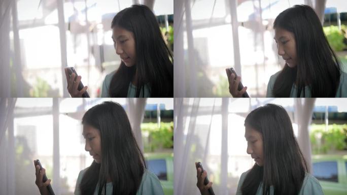 亚洲女孩使用智能手机，站在家里的窗户附近。
