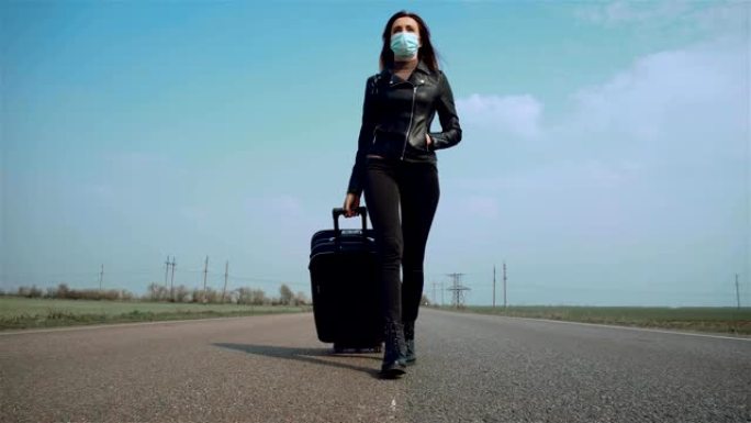 戴着口罩的独身女人走在路中间的中间地带，带着轮子上的手提箱