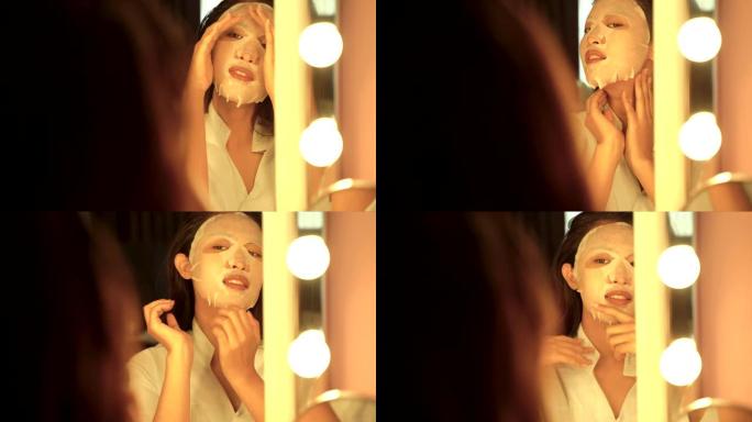 年轻的亚洲妇女正在她的脸上涂上面膜，看着她在浴室里的倒影