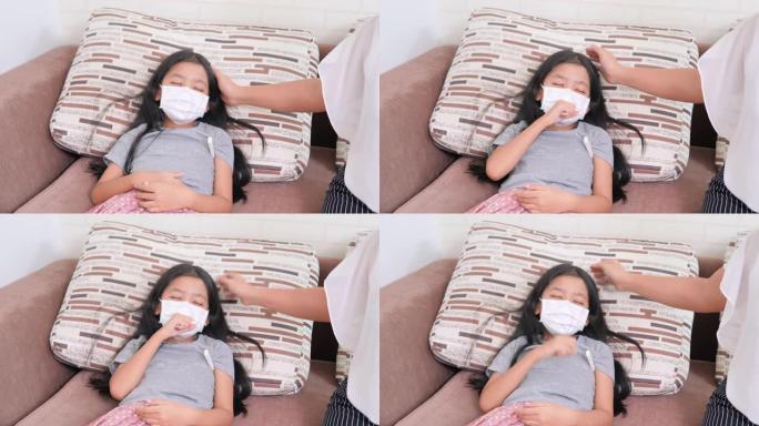 亚洲小女孩戴着外科口罩，躺在咳嗽的沙发上。妇女使用儿童温度计。