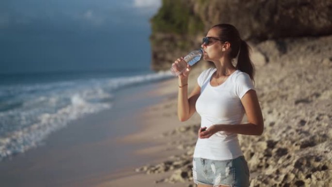 戴着太阳镜的美丽年轻女子在海滩上喝瓶子里的清水