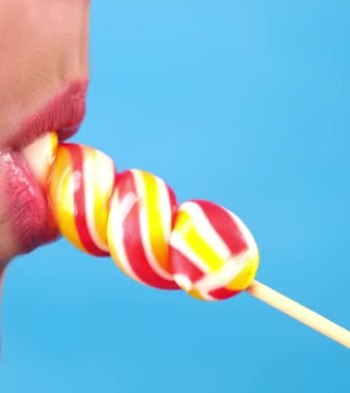 垂直特写，女性性感的嘴唇用红色唇膏吮吸大螺旋长方形糖果棒棒糖。复制空间。蓝色背景