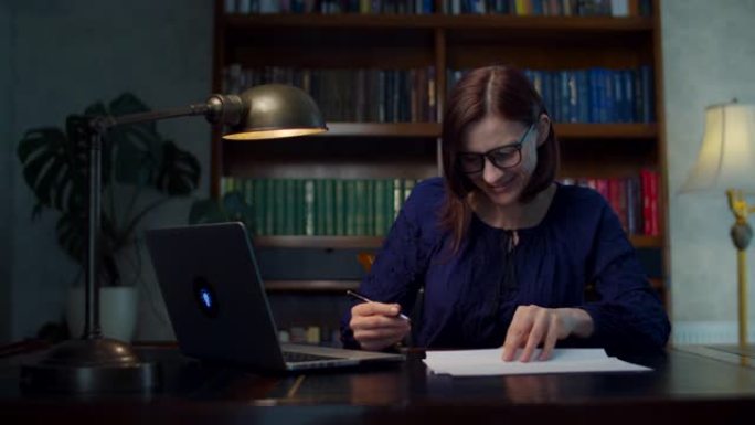 30岁戴眼镜的快乐黑发女人在家工作，坐在带书柜的木桌前签署纸质文件。微笑的女人在家工作。