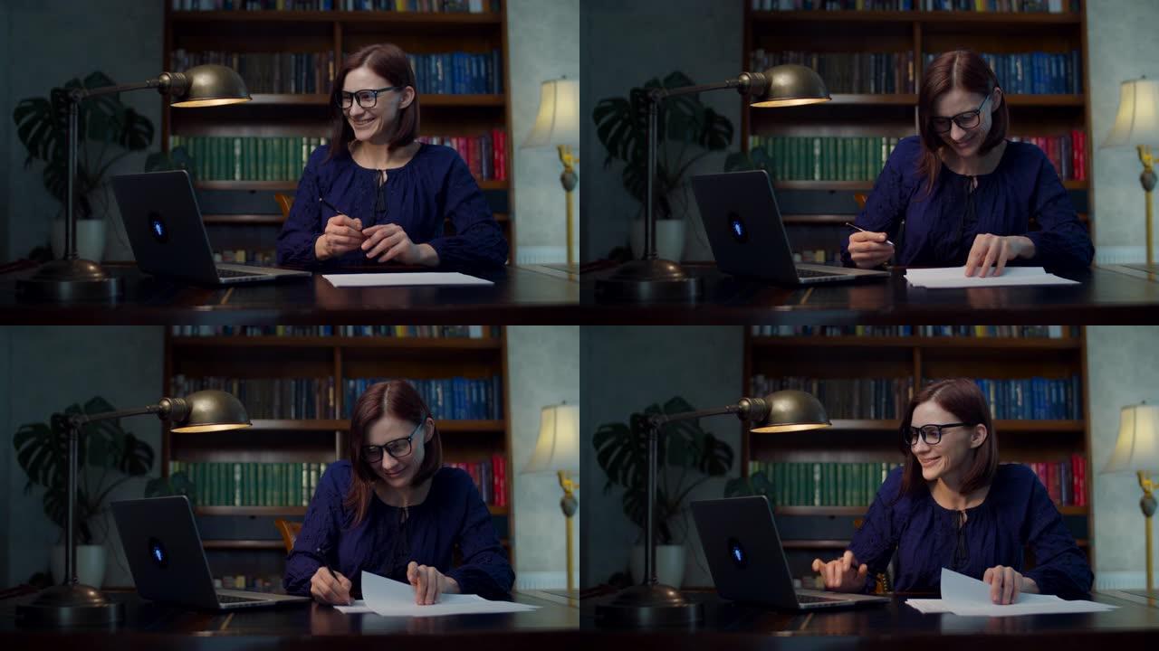 30岁戴眼镜的快乐黑发女人在家工作，坐在带书柜的木桌前签署纸质文件。微笑的女人在家工作。