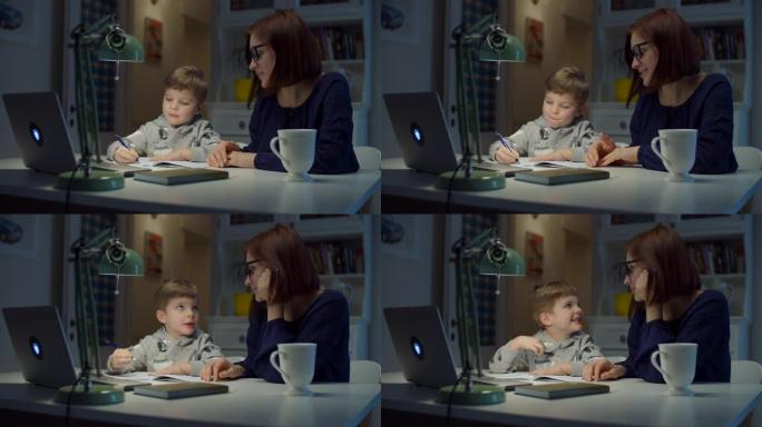 30多岁的年轻母亲在家与学龄前儿童的儿子一起做在线作业。戴眼镜的女人和小男孩用笔记本电脑，笔记本电脑