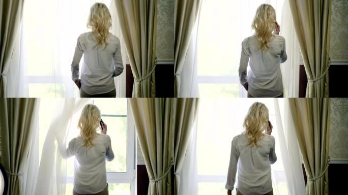 迷人的金发女人在手机上聊天，看着窗户。4K