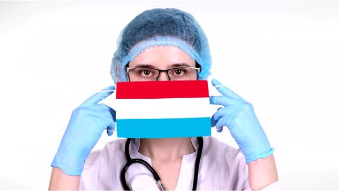 特写。戴眼镜的医生，蓝色医疗帽，手套手持带有卢森堡国旗的医用口罩。医生护理，冠状病毒期间国家保护，全