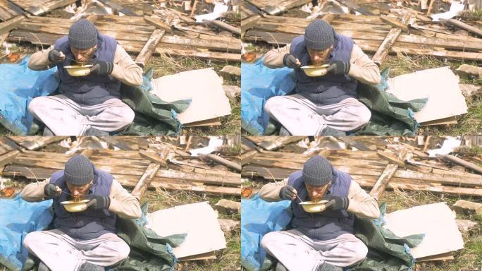 一个无家可归的人从废墟附近的盘子里吃汤，在疫情期间帮助贫困和饥饿的人