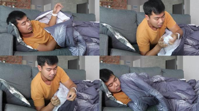 一名亚洲男子生病，在家中的沙发上咳嗽，他在冠状病毒流行期间使用温度计检查温度。