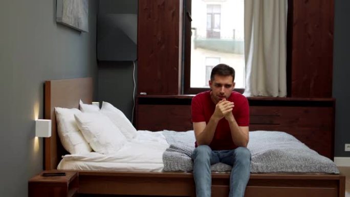 沮丧的年轻人坐在家里或酒店房间的床边，焦虑地揉手。患有药物滥用或精神障碍的紧张男子