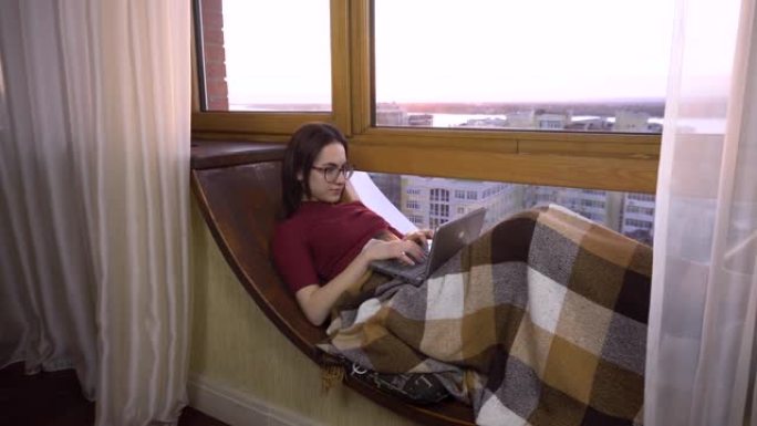 一名年轻女子正在笔记本电脑上打字。一个女孩躺在窗边的窗台上，手里拿着笔记本电脑。窗外是明亮的一天。