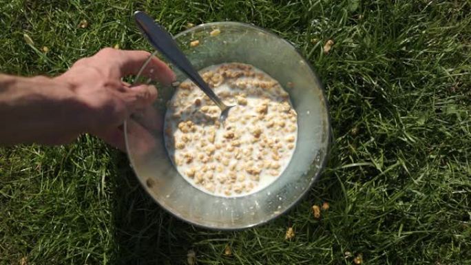 早餐准备好了。男人用手将牛奶填充牛奶的麦片脆麦片放在绿色草地草地上的透明碗中