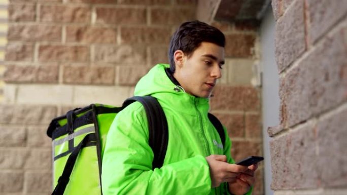 绿色制服的年轻男性食品快递员的侧视图，手机上有保温袋阅读订单，同时打电话给门铃并与客户交谈