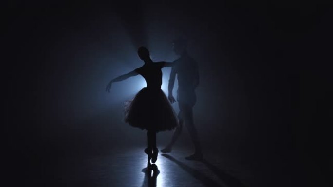 优雅的芭蕾舞演员和她的男搭档在黑暗中用泛光灯背光跳舞古典或现代芭蕾舞的元素。黑色背景上的烟雾情侣。艺