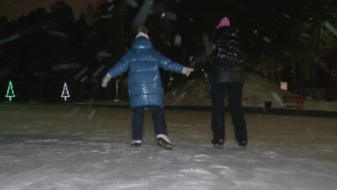 妈妈一起下雪时，女儿在冬季公园的夜间溜冰场上滑冰。母亲和少女在户外冬季溜冰场上骑着冰鞋时手牵手。