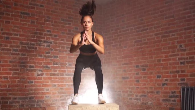 美丽的女性健美运动员在健身房表演蹲在健身盒上跳跃