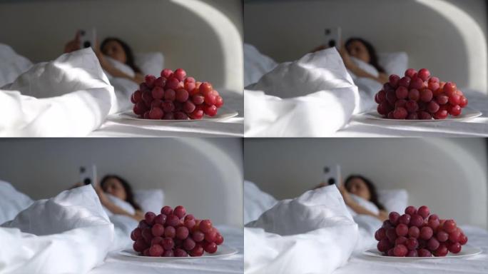 女人躺在床上拿着手机，阳光落在红葡萄盘子上