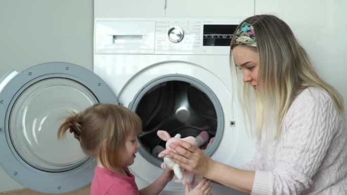 微笑的母亲带着3岁的小女儿做家务，把玩具放进洗衣机。女孩关闭舱门，打开家用电器。