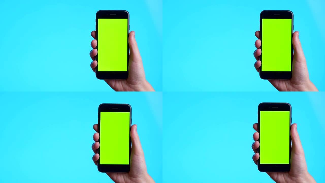 女人的手在蓝色背景上拿着带有绿色屏幕的智能手机。仅手的特写镜头