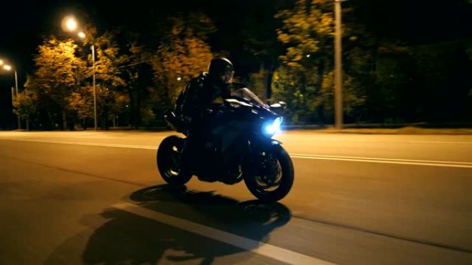 戴着头盔的年轻人在晚上的城市街道上快速骑现代黑色运动摩托车。摩托车手在夜间空旷的道路上骑摩托车。开车