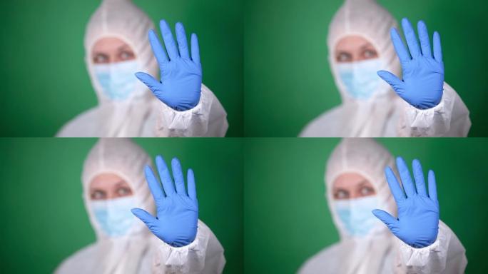 年轻的女孩医生穿着白色的抗瘟疫防护外套和蓝色的橡胶手套，在绿色背景下显示出隔离的停止手势