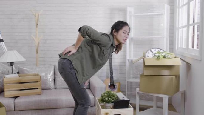 年轻的亚洲妇女背部疼痛，在新房客厅搬箱子。女孩在举起大纸箱纸板时伤了疼的腰。受伤不良姿势错误脊椎疼痛