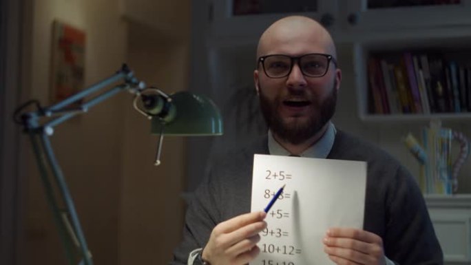 戴着眼镜的大胡子的30多岁男子展示着带有简单数学方程式的纸张，看着家里的相机。在线教育过程。老师说话