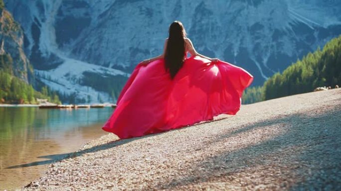 穿着红色连衣裙的艺术女性女王。梦幻长发丝绸面料飞风。慢动作女士没有脸地转过身来。自然山脉山脉的力量意