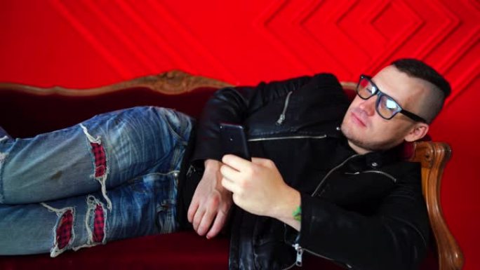 戴着眼镜的帅气小伙子坐在手机上靠着红墙的小沙发上。成年迷人的男性在手机上翻阅各种新闻，躺在扶手椅上