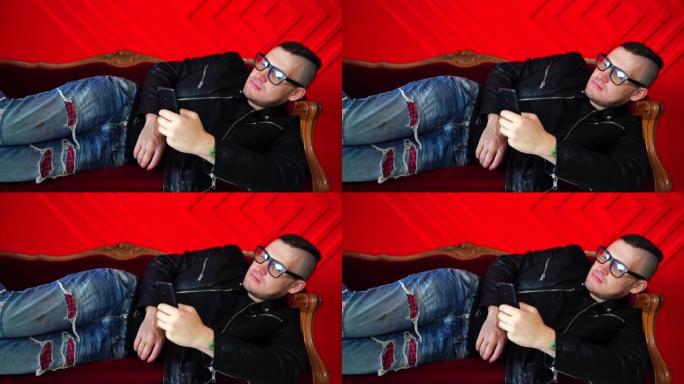 戴着眼镜的帅气小伙子坐在手机上靠着红墙的小沙发上。成年迷人的男性在手机上翻阅各种新闻，躺在扶手椅上