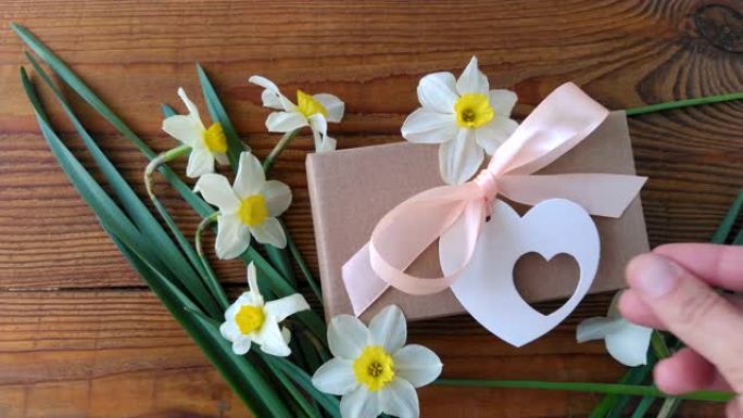 白色水仙花和棕色礼品盒，木质背景上带有粉红色丝带的贺卡4k视频。女性手在假期礼物周围放鲜花。3月8日