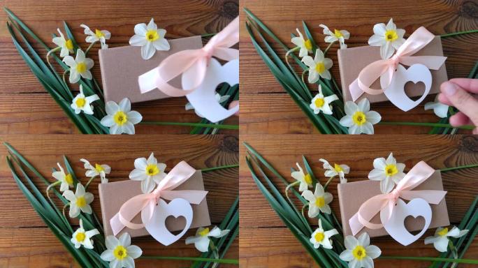 白色水仙花和棕色礼品盒，木质背景上带有粉红色丝带的贺卡4k视频。女性手在假期礼物周围放鲜花。3月8日