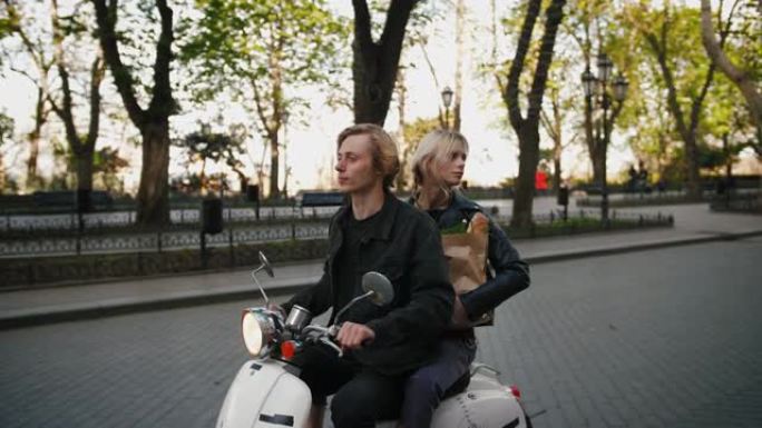 快乐的年轻时髦夫妇在旧城中心公园骑着一辆老式踏板车，带着杂货纸袋，跟踪镜头