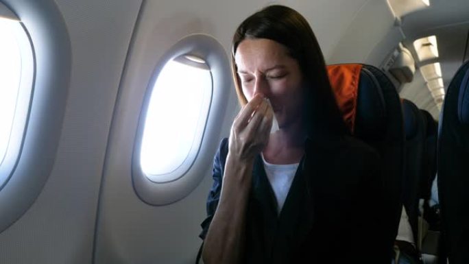 患有鼻炎，患有鼻涕的不健康女人。生病的女孩拿着纸巾吹鼻塞。她坐在飞机靠窗的乘客座位上。流感症状，慢性