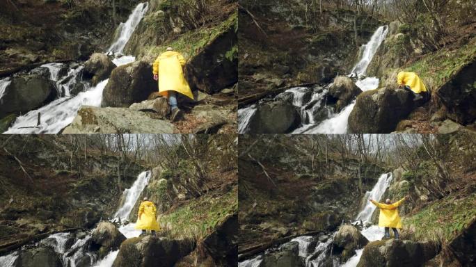 穿着黄色雨衣的年轻男性旅行者享受着美丽的瀑布。在山里徒步旅行。徒步旅行者奔向瀑布，举起双手。享受生活