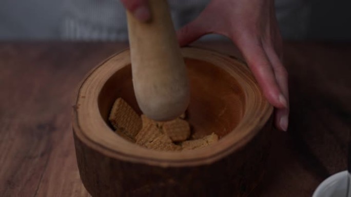 一个女人的手从一个装有饼干的白色碗中取出几块饼干，然后将它们放在木杵和研钵中，将其粉碎并磨成细粉。